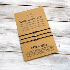 Op de afbeelding: Twee zwarte koordarmbanden met zilveren sterbedels en een kaart met de tekst "When We're Apart" en "Little Wishers". De kaart legt uit dat de armbanden bedoeld zijn voor vrienden om te dragen wanneer ze uit elkaar zijn en dat wanneer het koord breekt, een wens uitkomt.