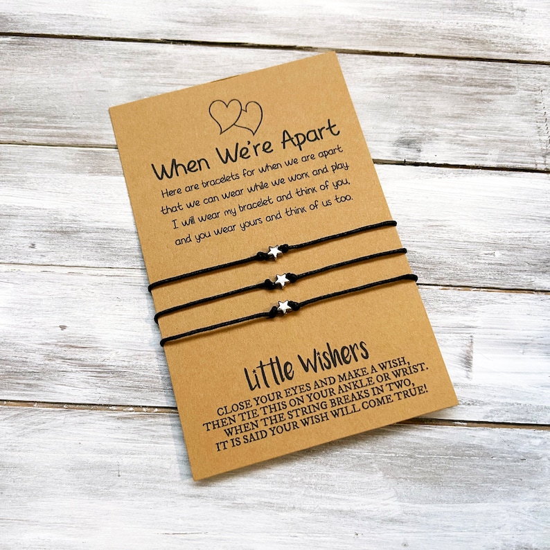 Op de afbeelding: Twee zwarte koordarmbanden met zilveren sterbedels en een kaart met de tekst "When We're Apart" en "Little Wishers". De kaart legt uit dat de armbanden bedoeld zijn voor vrienden om te dragen wanneer ze uit elkaar zijn en dat wanneer het koord breekt, een wens uitkomt.