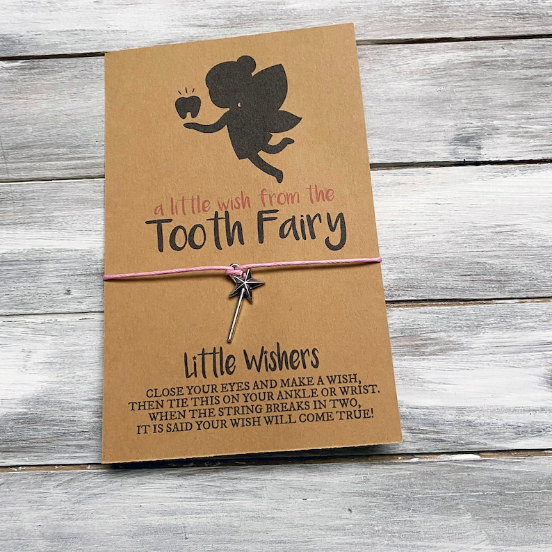 May include: A brown card with a silhouette of a tooth fairy holding a tooth. The card says "a little wish from the Tooth Fairy". A pink string with a silver star charm is attached to the card. The card also says "Little Wishers" and includes instructions on how to make a wish.