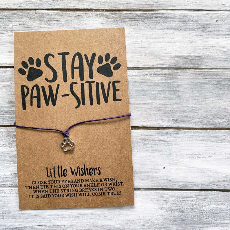 May include: A brown card with the text "STAY PAW-SITIVE" and a paw print charm attached to a purple string. The card also includes instructions for making a wish with the string bracelet.