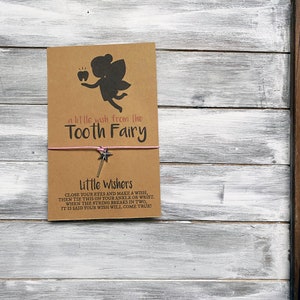 May include: A brown card with a pink string tied around it. The card has a silhouette of a tooth fairy with a tooth. The text on the card reads "a little wish from the Tooth Fairy" and "Little Wishers CLOSE YOUR EYES AND MAKE A WISH, THEN TIE THIS ON YOUR ANKLE OR WRIST. WHEN THE STRING BREAKS IN TWO, IT IS SAID YOUR WISH WILL COME TRUE!"
