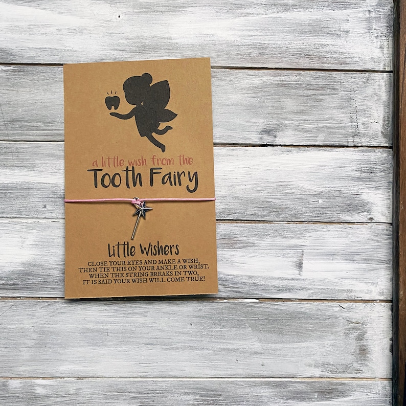 May include: A brown card with a pink string tied around it. The card has a silhouette of a tooth fairy with a tooth. The text on the card reads "a little wish from the Tooth Fairy" and "Little Wishers CLOSE YOUR EYES AND MAKE A WISH, THEN TIE THIS ON YOUR ANKLE OR WRIST. WHEN THE STRING BREAKS IN TWO, IT IS SAID YOUR WISH WILL COME TRUE!"