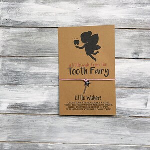 May include: A brown card with a silhouette of a tooth fairy holding a tooth. The card says "a little wish from the Tooth Fairy". A pink string with a silver star charm is attached to the card. The card also says "Little Wishers Close your eyes and make a wish, then tie this on your ankle or wrist. When the string breaks in two, it is said your wish will come true!"