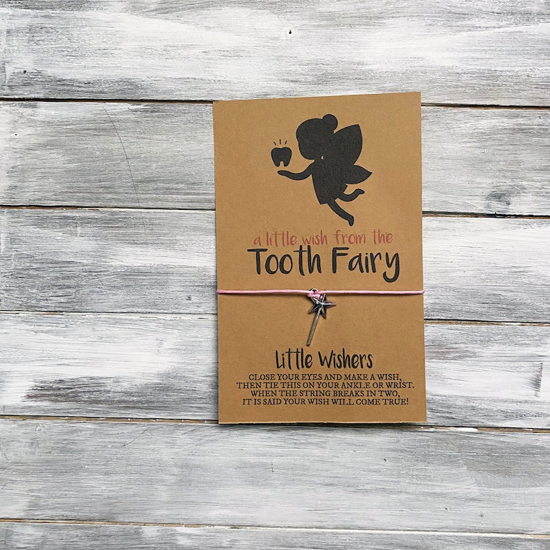 May include: A brown card with a silhouette of a tooth fairy holding a tooth. The card says "a little wish from the Tooth Fairy". A pink string with a silver star charm is attached to the card. The card also says "Little Wishers Close your eyes and make a wish, then tie this on your ankle or wrist. When the string breaks in two, it is said your wish will come true!"