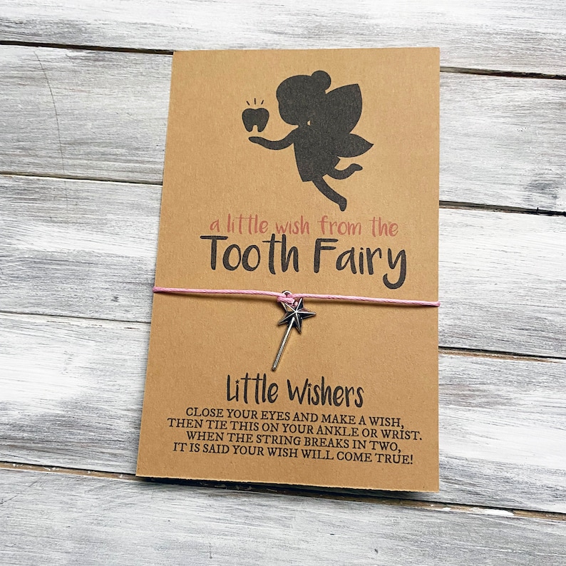 May include: A brown card with a silhouette of a tooth fairy holding a tooth. The card says "a little wish from the Tooth Fairy" and "Little Wishers".  The card is tied with a pink string and a silver star charm.