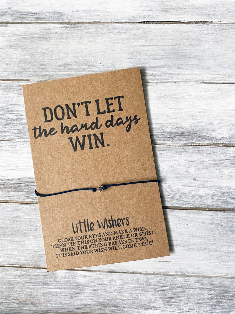 Puede incluir: Una tarjeta marr&oacute;n con el texto "DON'T LET the hard days WIN." y el texto "Little Wishers CLOSE YOUR EYES AND MAKE A WISH, THEN TIE THIS ON YOUR ANKLE OR WRIST. WHEN THE STRING BREAKS IN TWO, IT IS SAID YOUR WISH WILL COME TRUE!" Una pulsera de cuerda negra con un dije de estrella plateada est&aacute; atada alrededor de la tarjeta.