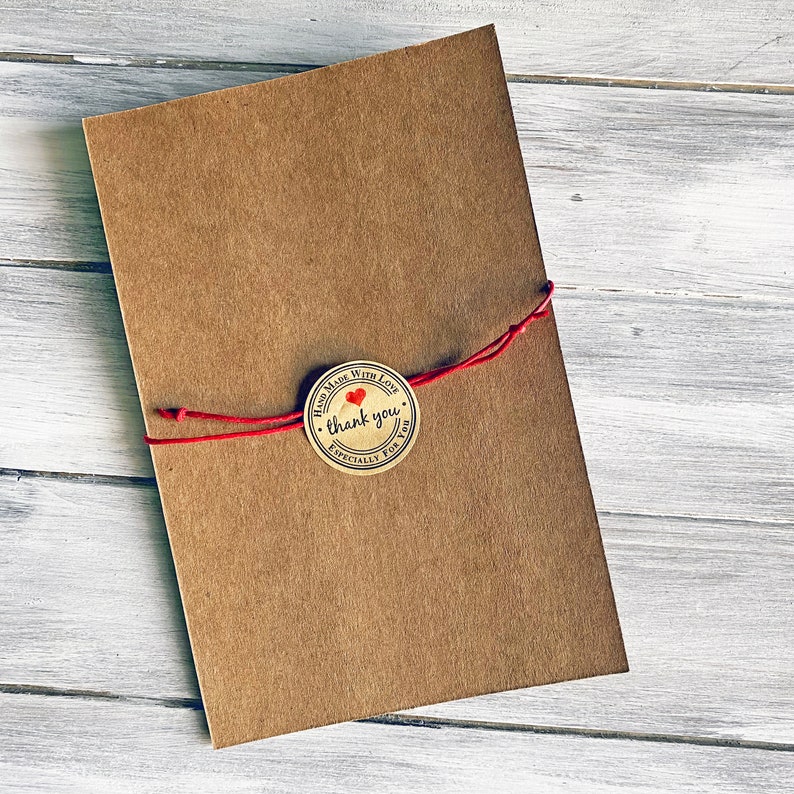 May include: A brown paper envelope tied with red string and a round, brown paper sticker with a red heart and the text "Hand Made With Love, thank you, Especially For You".