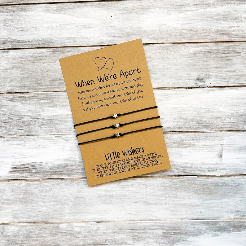 Op de afbeelding: Twee zwarte vriendschapsarmbanden met zilveren ster-bedels en een kaart met de tekst "When We're Apart" en "Little Wishers". De kaart legt uit dat de armbanden zijn voor wanneer je uit elkaar bent en dat je ze kunt dragen terwijl je werkt en speelt. Het legt ook uit dat wanneer de draad in twee&euml;n breekt, het gezegd wordt dat je wens uitkomt.