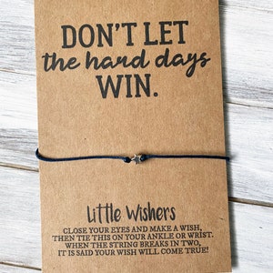 Puede incluir: Una tarjeta marr&oacute;n con el texto "DON'T LET the hard days WIN." y una pulsera de cuerda negra con un dije de estrella plateada. La tarjeta tambi&eacute;n incluye el texto "Little Wishers" e instrucciones para hacer un deseo con la pulsera.