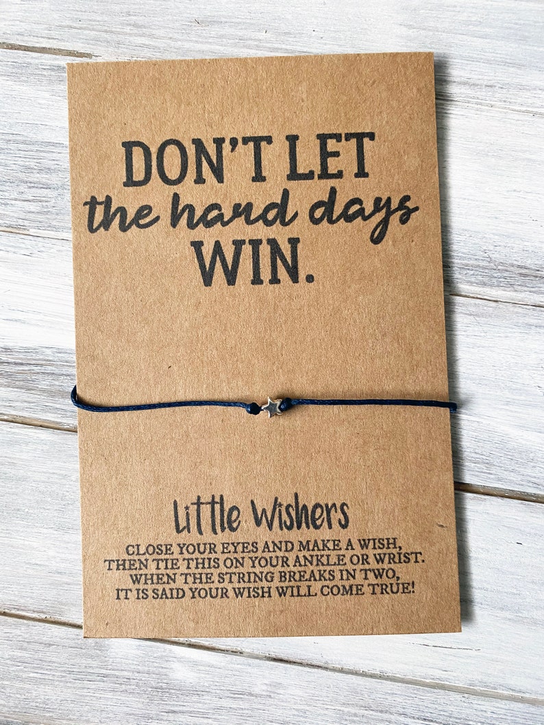 Puede incluir: Una tarjeta marr&oacute;n con el texto "DON'T LET the hard days WIN." y una pulsera de cuerda negra con un dije de estrella plateada. La tarjeta tambi&eacute;n incluye el texto "Little Wishers" e instrucciones para hacer un deseo con la pulsera.