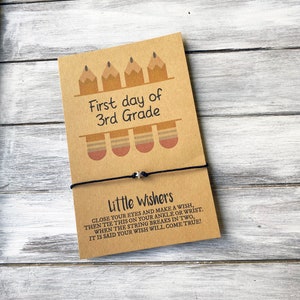 May include: A brown card with the text "First day of 3rd Grade" and a pencil illustration. A black string bracelet with a silver star charm is tied around the card. The card reads "Little Wishers Close your eyes and make a wish, then tie this on your ankle or wrist. When the string breaks in two, it is said your wish will come true!"