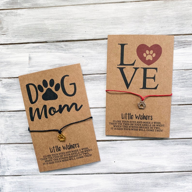 May include: Two brown cards with paw print charms attached to red and black strings. The cards have the text "Dog Mom" and "Love" with paw print designs. The cards also have instructions for making a wish with the string.