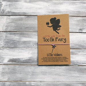 May include: A brown card with a silhouette of a tooth fairy holding a tooth. The card says "a little wish from the Tooth Fairy". Below the card is a pink string with a silver star charm attached to it. The text below the charm says "Little Wishers Close your eyes and make a wish, then tie this on your ankle or wrist. When the string breaks in two, it is said your wish will come true!"