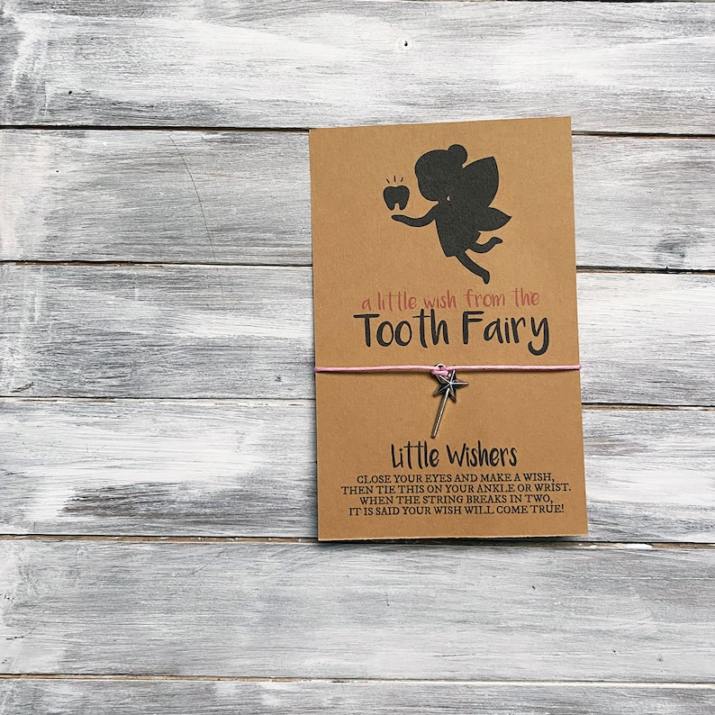 May include: A brown card with a silhouette of a tooth fairy holding a tooth. The card says "a little wish from the Tooth Fairy". Below the card is a pink string with a silver star charm attached to it. The text below the charm says "Little Wishers Close your eyes and make a wish, then tie this on your ankle or wrist. When the string breaks in two, it is said your wish will come true!"