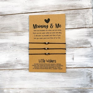 Puede incluir: Un conjunto de dos pulseras de cuerda negra con abalorios de estrella plateada. La tarjeta dice "Mommy & Me" y "Little Wishers". La tarjeta explica que las pulseras son para cuando estás lejos el uno del otro y que puedes llevarlas mientras trabajas y juegas. También dice que cierres los ojos y pidas un deseo, luego ata la pulsera a tu tobillo o muñeca. Cuando la cuerda se rompe en dos, se dice que tu deseo se hará realidad.