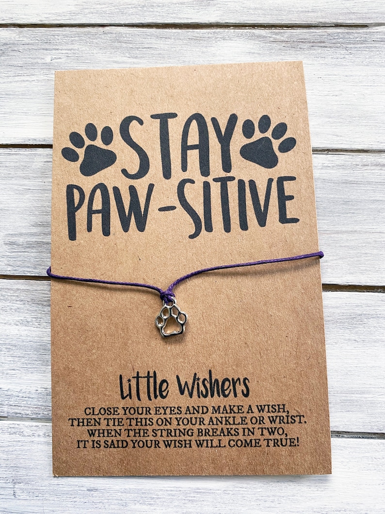 May include: A brown card with the text "STAY PAW-SITIVE" and a paw print charm attached to a purple string. The card also includes instructions for making a wish with the string bracelet.