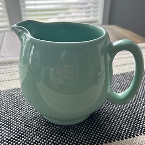 May include: A solid light mint green ceramic pitcher with a smooth, glossy finish. The pitcher features a rounded body, a curved handle on the right side, and a distinct pouring spout at the top rim, showcasing a simple and functional design.