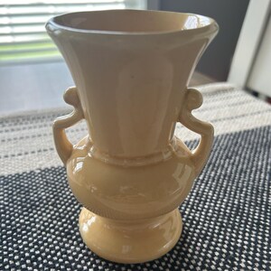 May include: A pale yellow ceramic vase with a glossy finish, featuring a wide flared rim, a rounded bulbous body, and two symmetrical curved handles on the sides. The vase stands on a circular pedestal base and has a smooth, solid-colored surface.