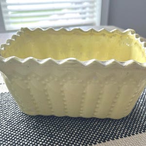 May include: A rectangular, pale yellow ceramic planter featuring a scalloped top edge and vertical rows of raised, beaded detailing along the exterior sides. The vessel has a smooth, glossy finish and an open, hollow interior for holding items.