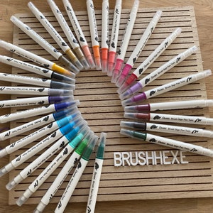 Könnte beinhalten: Ein Set mit 24 doppelseitigen Pinselstiften in verschiedenen Farben, darunter Schwarz, Blau, Grün, Rot und Gelb. Die Stifte sind in einem Kreis auf einer Holzoberfläche angeordnet, auf der der Text "BRUSHHEXE" aufgedruckt ist.