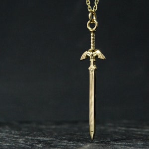 May include: A gold-colored necklace with a pendant shaped like a sword. The sword has a detailed design and a pointed tip.
