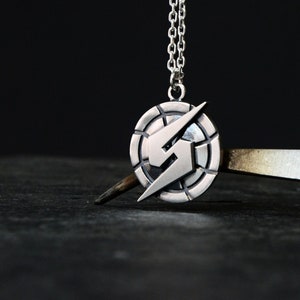 Puede incluir: Un collar con un colgante de plata que presenta un diseño de rayo estilizado dentro de un marco circular. El colgante cuelga de una cadena de plata.
