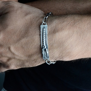May include: A silver bracelet with a textured rectangular charm featuring a stylized design of a trident.