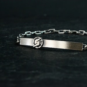 Puede incluir: Una pulsera de plata con un diseño de cadena de eslabones y una etiqueta rectangular con un símbolo de rayo estilizado.