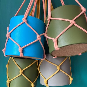 May include: Four hanging planters with different colored pots and macrame rope. The pots are blue, green, gray, and green. The rope is pink and yellow.