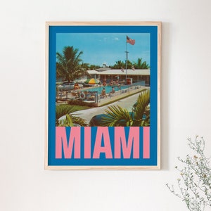 May include: Framed vintage-style poster featuring a pool scene in Miami. The image has a blue border and the word "MIAMI" in pink block letters at the bottom. The scene includes palm trees, a pool, and an American flag.