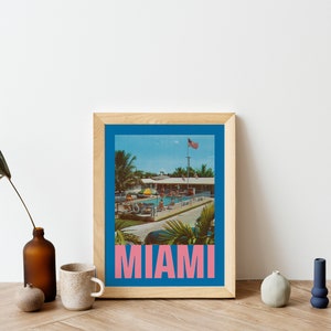 May include: A framed print featuring a vintage-style image of a pool scene in Miami. The image has a blue border and the word "MIAMI" in pink block letters at the bottom. The print is in a light wood frame.