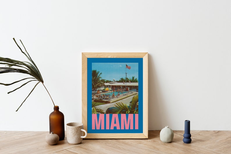May include: A framed print featuring a vintage-style image of a pool scene in Miami. The image has a blue border and the word "MIAMI" in pink block letters at the bottom. The print is in a light wood frame.