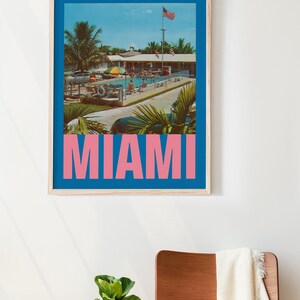 May include: A framed print featuring a vintage-style image of a pool scene in Miami. The image has a blue border and the word "MIAMI" in pink block letters. The scene includes palm trees, a pool, and people.