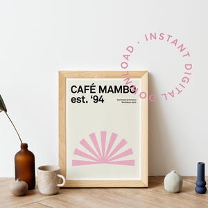 May include: A framed print with a pink sunburst design and the text "CAFÉ MAMBO est. '94". The print is in a wooden frame and is sitting on a wooden table with a vase, a mug, and other decorative items.