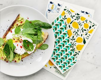 Puede incluir: Tostada de aguacate con huevo escalfado y espinacas en un plato, junto a paños de limpieza estampados. Los paños presentan diseños de limones y hojas en amarillo, verde y azul.