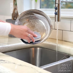 May include: A person washes a stainless steel pan with a gray and white cleaning sponge in a kitchen sink. The sink is made of stainless steel and has a white tiled backsplash. Water is running from the faucet.