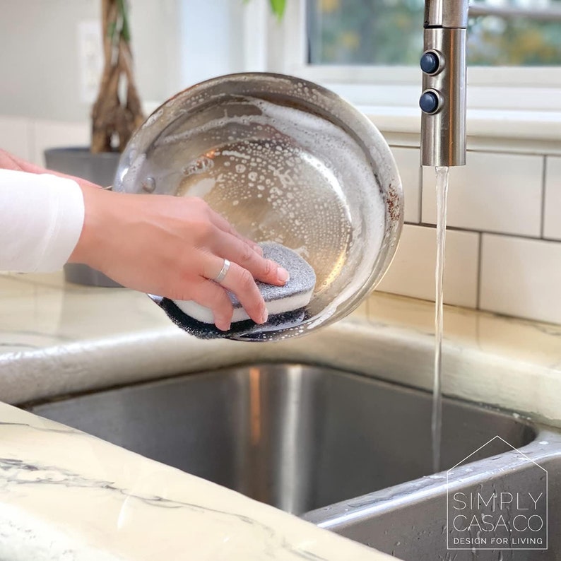 May include: A person washes a stainless steel pan with a gray and white cleaning sponge in a kitchen sink. The sink is made of stainless steel and has a white tiled backsplash. Water is running from the faucet.