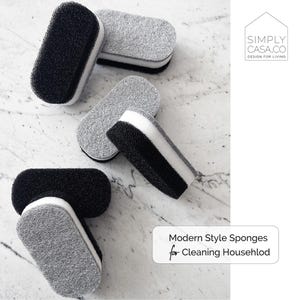May include: Four gray and black cleaning sponges with a white center. The sponges are arranged on a white marble surface. The text "Modern Style Sponges for Cleaning Household" is in the lower right corner of the image. The text "SIMPLY CASA.CO DESIGN FOR LIVING" is in the upper right corner of the image.