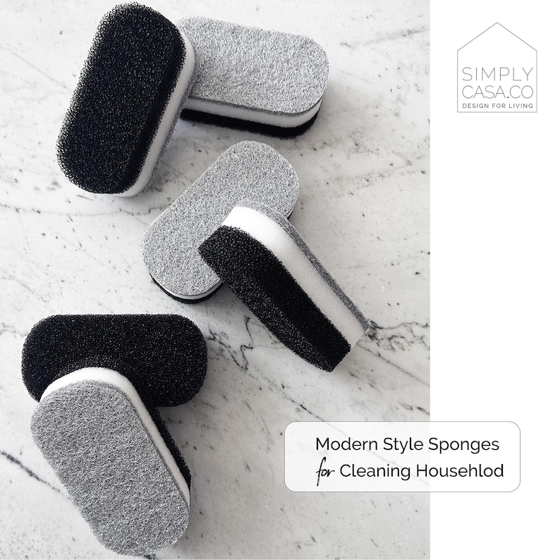 May include: Four gray and black cleaning sponges with a white center. The sponges are arranged on a white marble surface. The text "Modern Style Sponges for Cleaning Household" is in the lower right corner of the image. The text "SIMPLY CASA.CO DESIGN FOR LIVING" is in the upper right corner of the image.