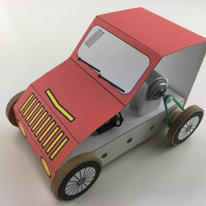 May include: A red cardboard car with a motor and a green belt. The car has a yellow and black grille and a white windscreen. The car is on a white surface.