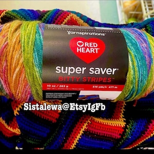 May include: A skein of Red Heart Super Saver Bitty Stripes yarn in a rainbow colorway, featuring a black label with the text Yarnspirations, Red Heart, super saver, BITTY STRIPES, 10 oz / 283 g, and 515 yds/v 471 m, resting on a crocheted blanket.