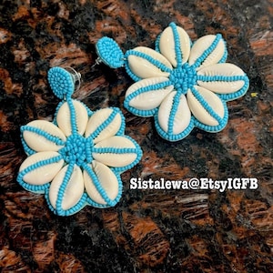 May include: A pair of flower-shaped earrings featuring white cowrie shells arranged in a circular pattern, accented with bright turquoise seed beads. The center and edges of each petal are lined with beads. Text reads Sistalewa@EtsyIGFB.