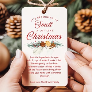 May include: A cream-colored Christmas-themed gift tag with the text "It's beginning to smell a lot like Christmas." The tag includes a recipe and is tied with twine. The background features Christmas ornaments and pine cones.