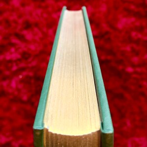 May include: Close-up of the spine of a hardback book with a green cover and gold-colored edges. The book is lying on a red surface.