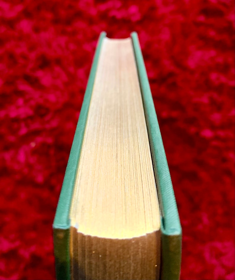 May include: Close-up of the spine of a hardback book with a green cover and gold-colored edges. The book is lying on a red surface.