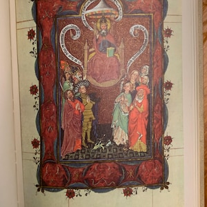 A medieval illustration of a religious scene. The central figure is a crowned figure seated on a throne, surrounded by other figures. The illustration is framed with a red and blue border with floral designs and text in a script language. The text reads 'Deus rex et dominus noster' and 'rex regum et dominus dominantium'.