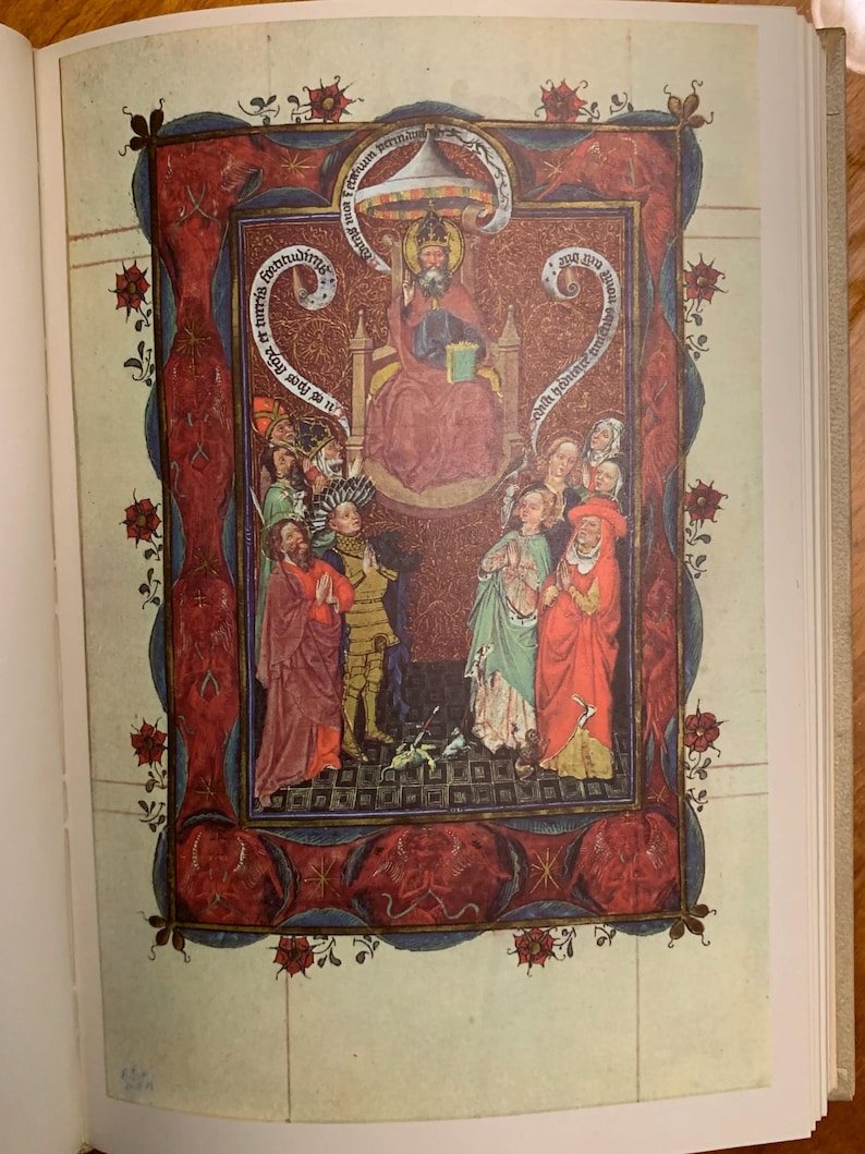 A medieval illustration of a religious scene. The central figure is a crowned figure seated on a throne, surrounded by other figures. The illustration is framed with a red and blue border with floral designs and text in a script language. The text reads 'Deus rex et dominus noster' and 'rex regum et dominus dominantium'.