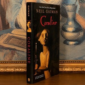 May include: A hardcover book titled Coraline by Neil Gaiman, featuring a dark cover illustration of a figure holding a lit candle. The spine displays the author's name and the title in red text against a black background, with a small portrait.