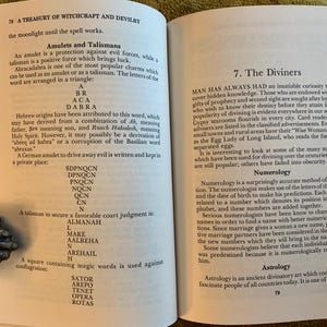 Puede incluir: P&aacute;ginas de un libro abierto con texto sobre brujer&iacute;a y adivinaci&oacute;n. El texto incluye secciones sobre amuletos, talismanes, numerolog&iacute;a y astrolog&iacute;a. El libro est&aacute; abierto por sujetalibros decorativos de metal.