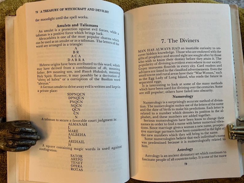 Puede incluir: P&aacute;ginas de un libro abierto con texto sobre brujer&iacute;a y adivinaci&oacute;n. El texto incluye secciones sobre amuletos, talismanes, numerolog&iacute;a y astrolog&iacute;a. El libro est&aacute; abierto por sujetalibros decorativos de metal.